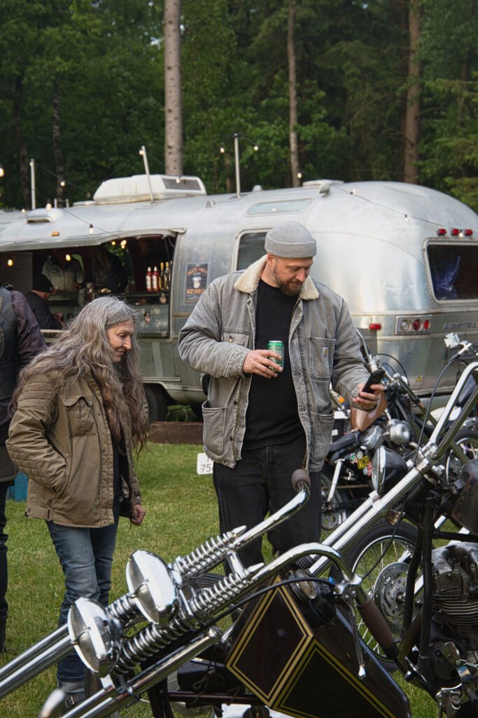 Ein Mann und eine Frau stehen bei einer Outdoor-Veranstaltung neben Custom-Motorrädern. Der Mann hält ein Getränk in der Hand und schaut auf sein Telefon. Ein Airstream-Anhänger und Bäume sind im Hintergrund zu sehen. Beide tragen Jacken; es scheint kühles Wetter zu sein.