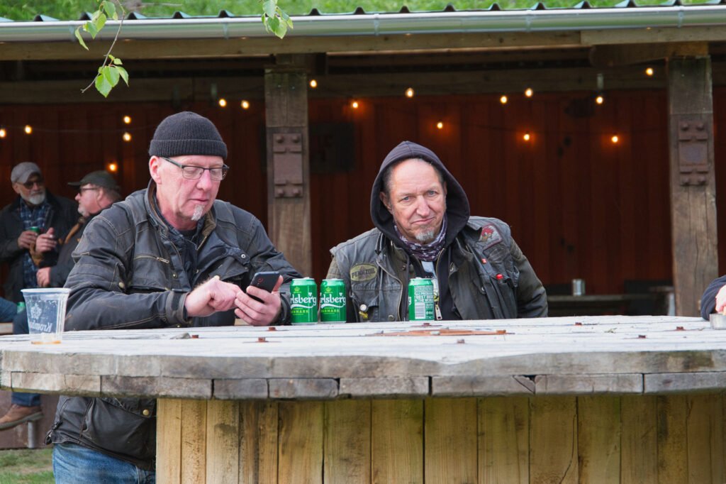 Zwei Männer in Jacken und Hüten sitzen an einem runden Holztisch im Freien, vor ihnen stehen Dosen mit Bier. Ein Mann telefoniert, während der andere in die Kamera schaut. Im Hintergrund sind Lichterketten und anderes zu sehen.