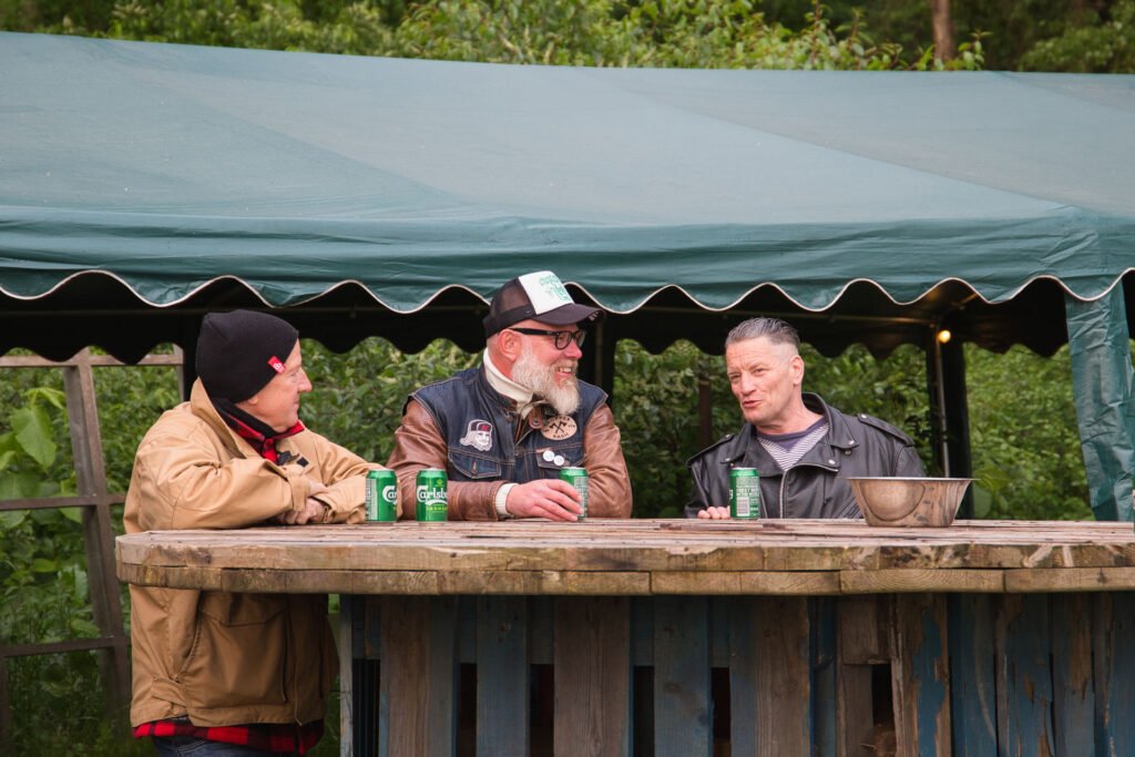 Drei Männer sitzen an einer rustikalen Bar im Freien, unterhalten sich und trinken Dosenbier. Sie tragen legere Jacken und Hüte, im Hintergrund stehen Bäume und ein grünes Zeltdach.