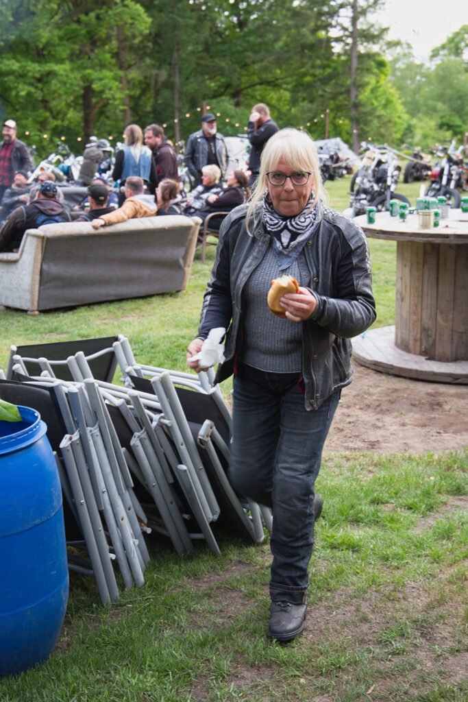 Eine Frau mit langen blonden Haaren, Brille und Lederjacke hält ein Sandwich und eine Serviette in der Hand. Sie geht im Freien bei einer Versammlung mit Menschen, Motorrädern und Klappstühlen im Hintergrund spazieren.