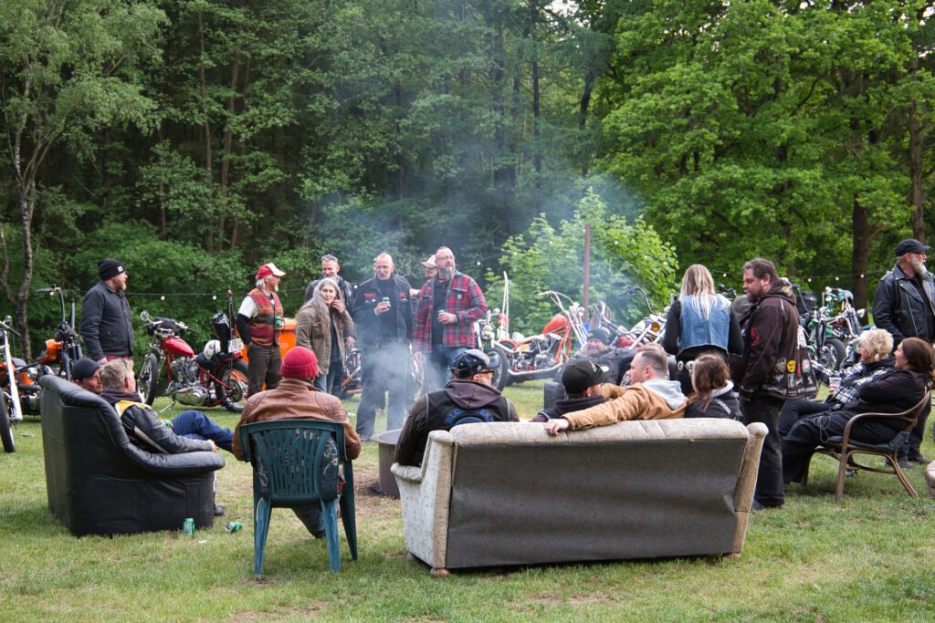 Eine Gruppe von Menschen versammelt sich um eine Feuerstelle und sitzt auf Sofas und Stühlen im Freien, während im Hintergrund Motorräder geparkt sind und Bäume die Gegend umgeben.