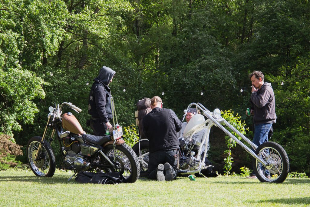 Vier Personen versammeln sich im Freien in der Nähe von zwei Custom-Motorrädern auf einer Wiese, mit Bäumen im Hintergrund. Eine Person sitzt auf einem Chopper, während die anderen in der Nähe stehen oder knien und sich unterhalten.