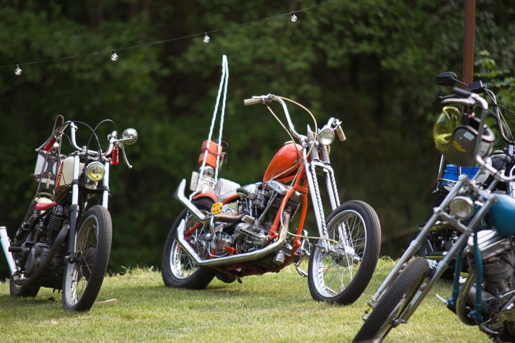 Mehrere Custom-Motorräder sind auf einer Wiese im Freien geparkt. Der orangefarbene Chopper in der Mitte hat einen hohen Lenker und eine Sissy Bar. Im Hintergrund sind Bäume und Lichterketten zu sehen.