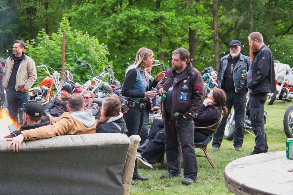 Eine Gruppe von Menschen in Biker-Kleidung trifft sich im Freien in der Nähe von Motorrädern, einige sitzen auf Sofas und Stühlen, andere stehen und unterhalten sich. Im Hintergrund stehen Bäume und Grünpflanzen, die eine lockere Atmosphäre schaffen.