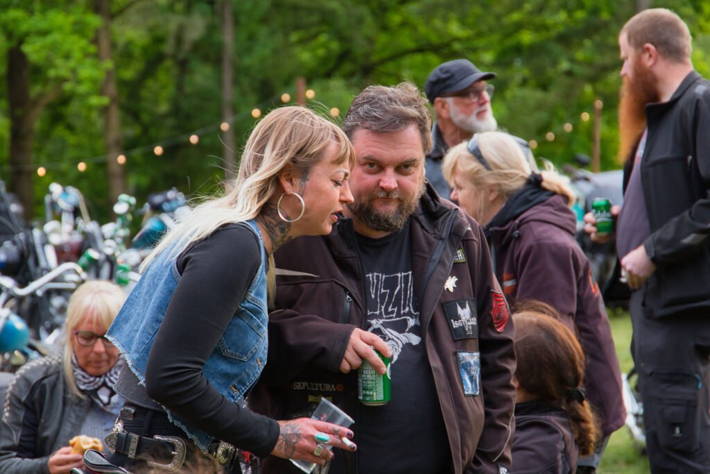 Eine Frau beugt sich vor, um mit einem Mann zu sprechen, der eine Dose bei einem Treffen im Freien hält. Im Hintergrund sind andere Menschen und Motorräder zu sehen, mit Lichterketten und grünen Bäumen ringsum.