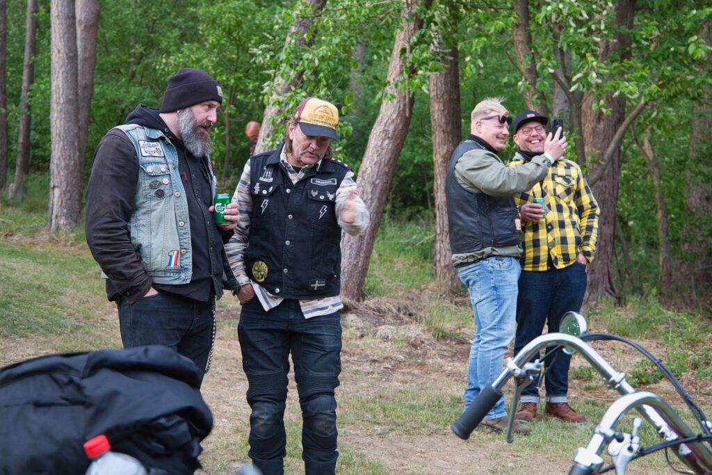 Vier Männer stehen im Freien in der Nähe von Bäumen, zwei davon im Vordergrund mit Motorradwesten und Hüten, und unterhalten sich. Zwei im Hintergrund unterhalten sich und lachen, einer gestikuliert energisch. Vorne ist teilweise ein Motorrad zu sehen.