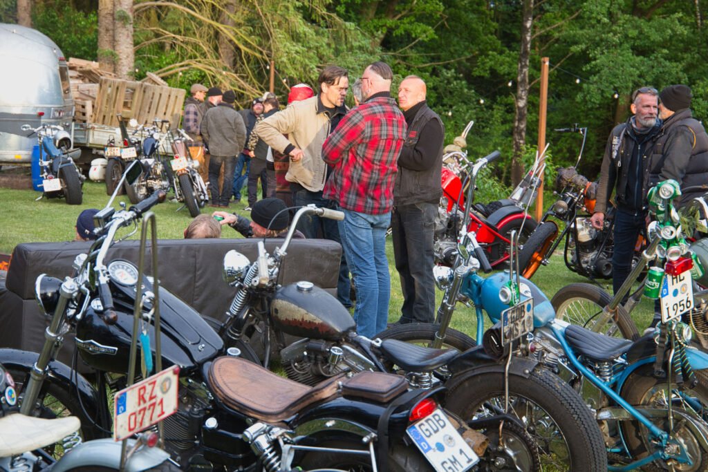 Eine Gruppe von Menschen unterhält sich und versammelt sich im Freien zwischen mehreren geparkten Oldtimer-Motorrädern auf einer Wiese, umgeben von Bäumen und einem Anhänger im Hintergrund.