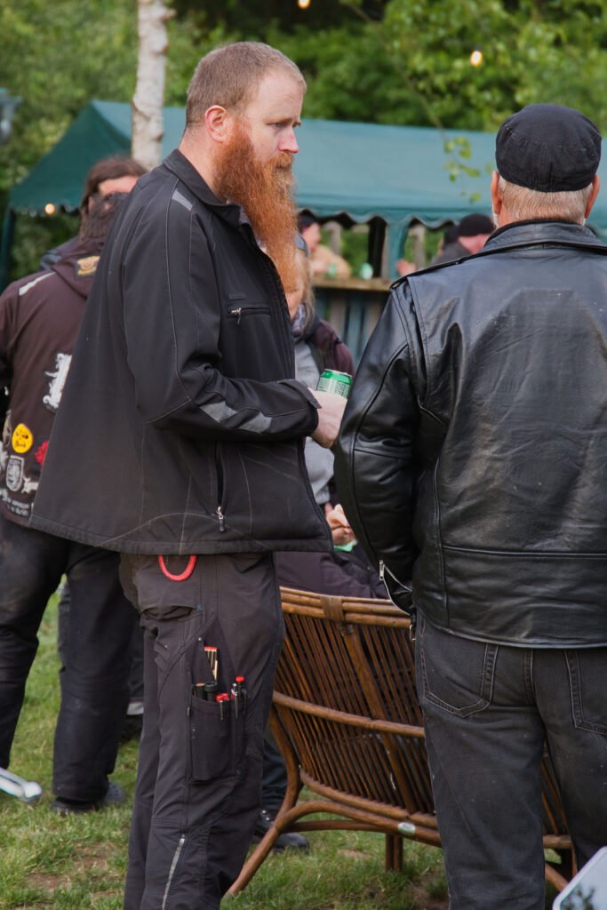 Ein Mann mit langem rotem Bart und schwarzer Jacke unterhält sich mit einem anderen Mann in schwarzer Lederjacke und Mütze bei einem Treffen im Freien, wobei im Hintergrund Menschen und ein grünes Zelt zu sehen sind.