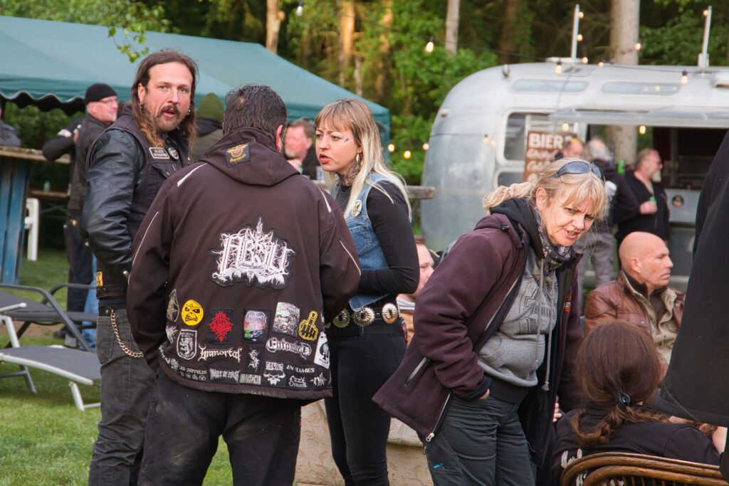 Eine Gruppe von Menschen in Bikerjacken und Kapuzenpullovern unterhält sich im Freien in der Nähe eines Airstream-Anhängers und eines grünen Vordachs, mit Bäumen und anderen Menschen im Hintergrund.
