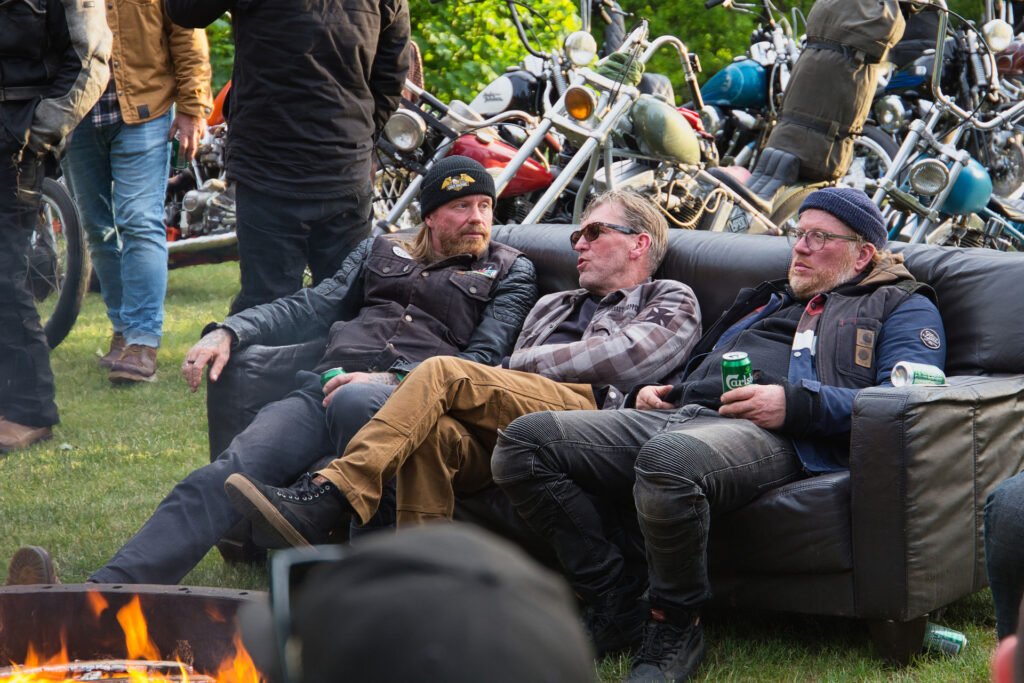 Drei Männer in legeren Jacken und Hüten sitzen auf einem schwarzen Sofa im Freien und halten Getränkedosen in der Hand, während hinter ihnen Motorräder geparkt sind und im Vordergrund ein Lagerfeuer brennt. Andere stehen und mischen sich in den Hintergrund.