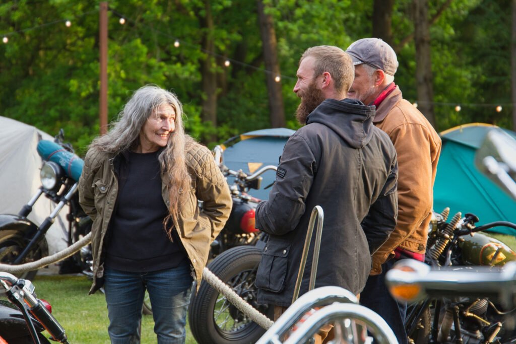 Drei Personen stehen und unterhalten sich im Freien neben Motorrädern und Zelten, umgeben von Bäumen und Lichterketten. Eine Frau mit langen grauen Haaren lächelt, während zwei Männer in Jacken zuhören und lachen.