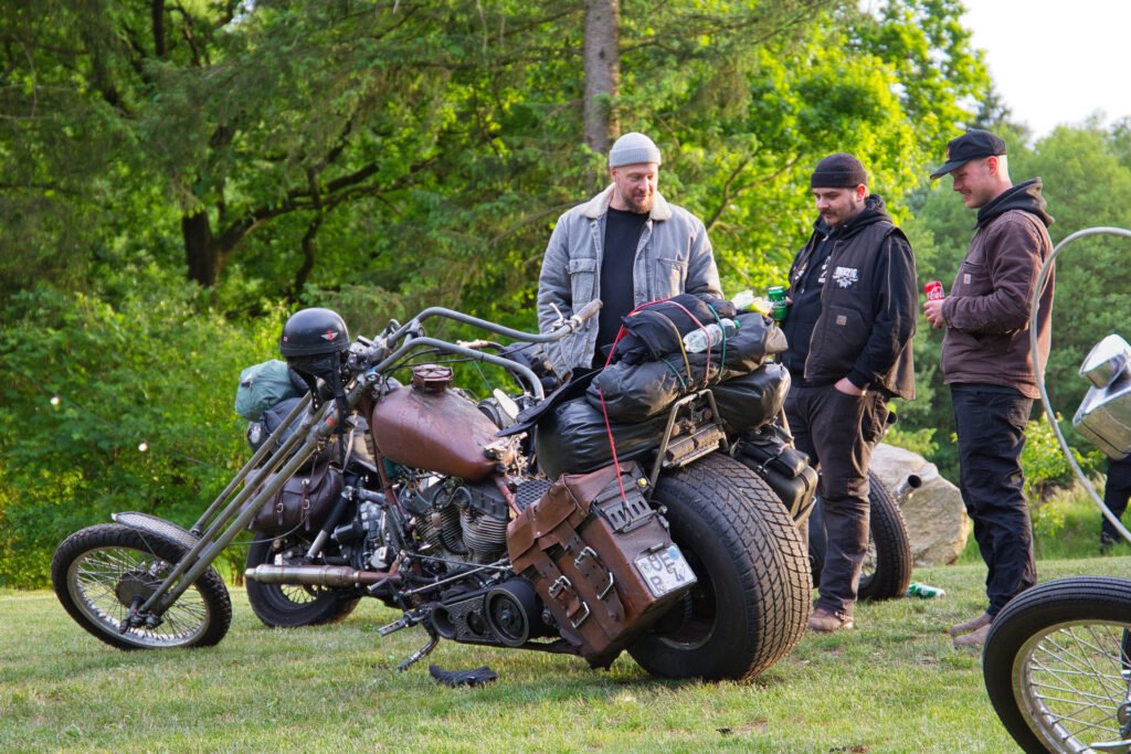Drei Männer stehen im Gras, lächeln und unterhalten sich in der Nähe eines geparkten, maßgeschneiderten Chopper-Motorrads, das mit Ausrüstung beladen ist. Im Hintergrund sind Bäume und Grünflächen zu sehen.