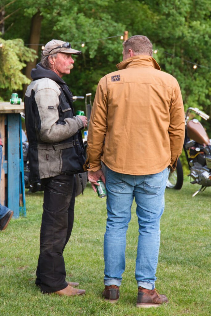 Zwei Männer stehen im Freien im Gras und unterhalten sich. Der eine trägt eine schwarze Jacke und Mütze und hält eine Dose in der Hand, der andere hat eine hellbraune Jacke und Jeans an und hält ebenfalls eine Dose in der Hand. Im Hintergrund sind Motorräder und Bäume zu sehen.