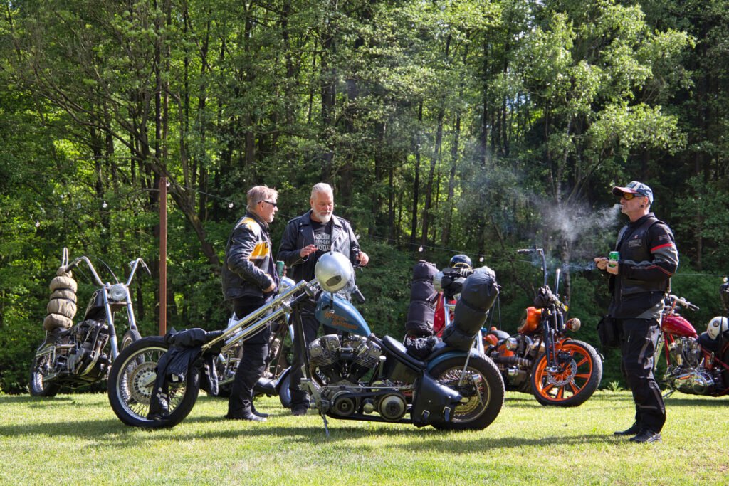 Drei Männer in Motorradkleidung stehen und unterhalten sich neben geparkten Motorrädern im Chopper-Stil auf einer Wiese, die von Bäumen umgeben ist. Ein Mann raucht. Auf den Motorrädern sind Motorradkleidung und Rucksäcke zu sehen.