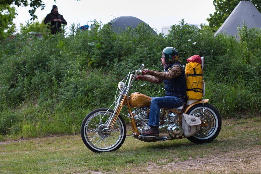 Eine Person, die einen Helm und eine Lederjacke trägt, fährt auf einem Motorrad im Vintage-Stil mit einem gelben Campingrucksack auf dem Rücken auf einer Wiese in der Nähe von Büschen und Zelten.