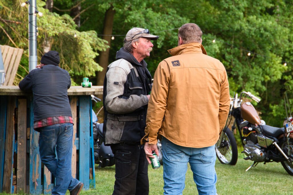 Drei Männer bei einem Treffen im Freien in der Nähe einer hölzernen Bar; zwei stehen im Gespräch, einer in einer hellbraunen Jacke, der andere in einer grau-schwarzen Jacke, während ein weiterer Mann an der Bar lehnt. Im Hintergrund sind Motorräder und Bäume zu sehen.