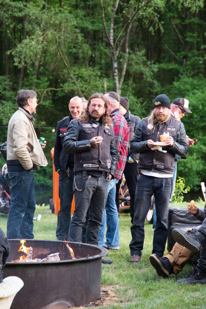 Eine Gruppe von Menschen in Bikerwesten und -jacken steht um eine Feuerstelle im Freien, mit Bäumen und Grün im Hintergrund, und unterhält sich. Einige halten Essen und Getränke in der Hand und genießen das entspannte Beisammensein.
