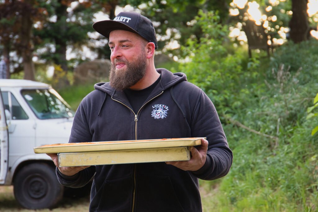 Ein bärtiger Mann mit schwarzem Kapuzenpulli und Mütze steht im Freien und hält ein großes Tablett. Im Hintergrund sind Grünpflanzen und ein weißer Lieferwagen zu sehen. Der Mann lächelt und scheint bei einer zwanglosen Zusammenkunft im Freien zu sein.