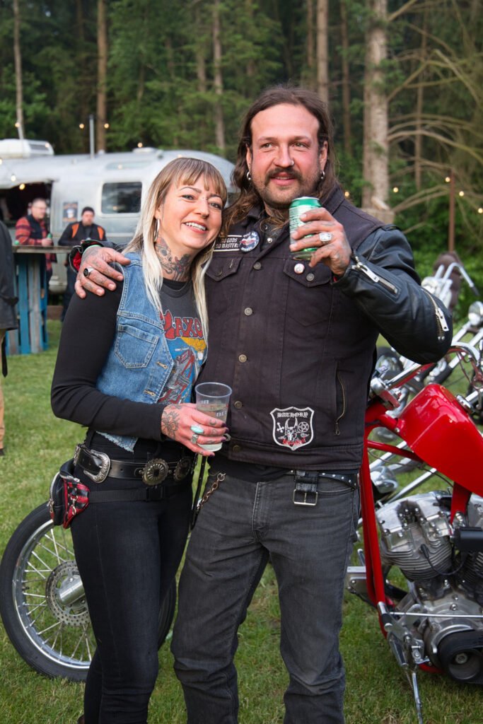 Ein lächelndes Paar mit Tätowierungen und Bikerwesten steht im Freien dicht beieinander und hält Getränke in der Hand. Sie stehen neben einem roten Motorrad mit Bäumen und Menschen im Hintergrund, was eine Zusammenkunft oder ein Fest zu sein scheint.