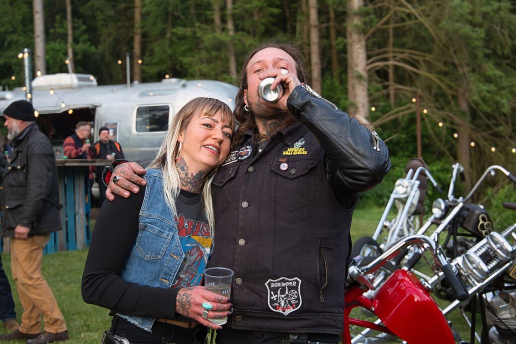 Eine lächelnde Frau mit Tätowierungen umarmt einen tätowierten Mann, der bei einer Veranstaltung im Freien mit Motorrädern, Bäumen, Lichterketten und einem silbernen Anhänger im Hintergrund aus einer Dose trinkt.