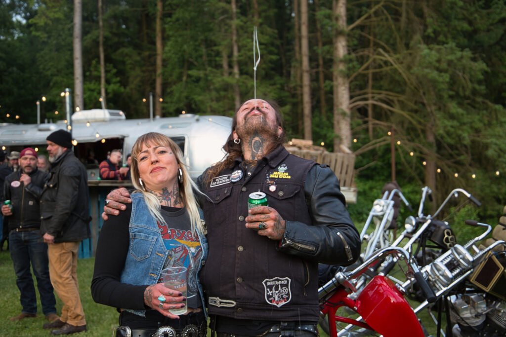 Eine Frau und ein Mann stehen zusammen bei einem Treffen im Freien mit Motorrädern und Menschen im Hintergrund; der Mann neigt seinen Kopf nach hinten und spuckt einen Strom von Flüssigkeit in die Luft.