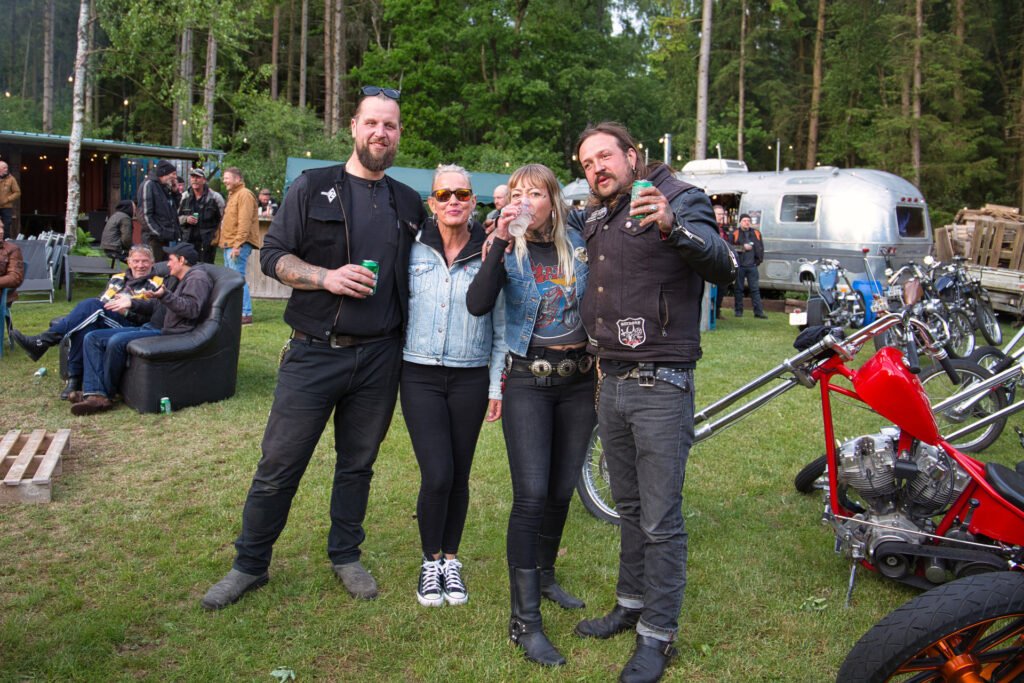 Vier Personen in legerer Biker-Kleidung stehen im Freien zusammen, lächeln und halten Getränke in der Hand, mit Motorrädern und einem silbernen Anhänger im Hintergrund bei einem Treffen im Wald. Andere Leute entspannen sich in der Nähe auf Stühlen.