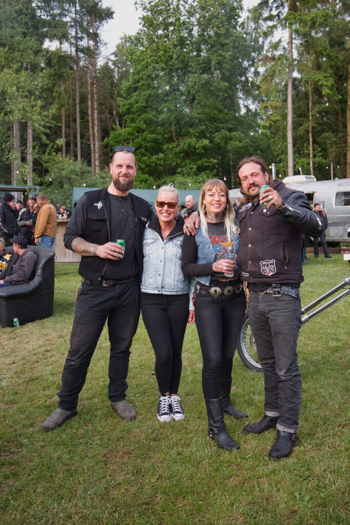 Vier Personen stehen lächelnd bei einer Veranstaltung im Freien zusammen, umgeben von Bäumen und Wohnmobilen. Zwei von ihnen halten Getränke in der Hand, und alle tragen Freizeit- oder Biker-Kleidung mit Jacken und Sonnenbrillen.