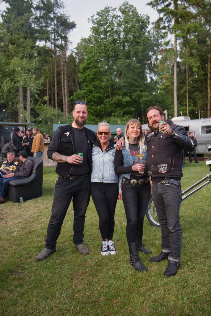 Vier Personen stehen zusammen im Freien auf einer Wiese, lächeln und posieren für ein Foto bei einer Veranstaltung. Sie tragen legere Kleidung im Biker-Stil, halten Getränke in der Hand und haben Bäume und andere Menschen im Hintergrund.