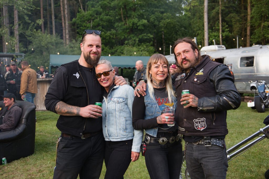 Vier Personen stehen bei einer Zusammenkunft im Freien zusammen und lächeln in die Kamera. Sie tragen Kleidung im Biker-Stil und halten Getränke in der Hand. Im Hintergrund sind Bäume und Menschen zu sehen, in der Nähe befinden sich ein Wohnmobil und Veranstaltungszelte.