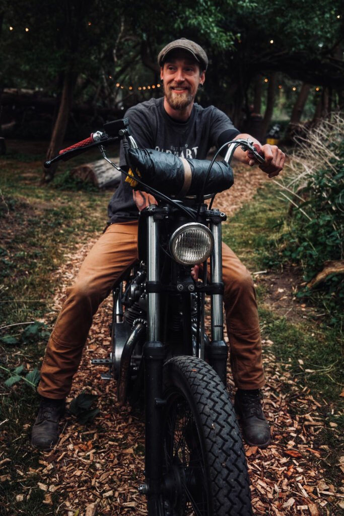 Ein Mann mit Bart und Mütze lächelt, während er auf einem schwarzen Motorrad auf einem mit Holzspänen bedeckten Waldweg sitzt, der von Grünpflanzen und Lichterketten umgeben ist.