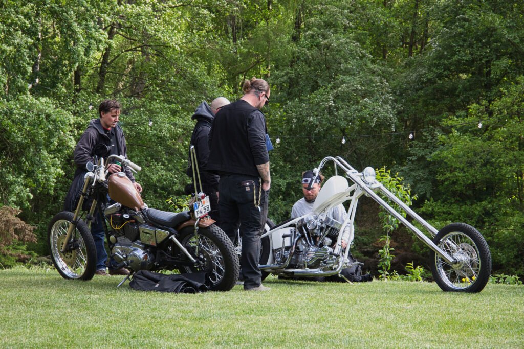 Vier Personen stehen und sitzen neben zwei Custom-Chopper-Motorrädern auf einer von Bäumen umgebenen Wiese, während im Hintergrund Lichterketten hängen.