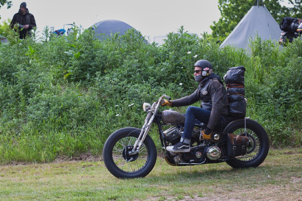 Ein Mann mit Helm und Handschuhen fährt auf einem verpackten Motorrad im Chopper-Stil an grünen Büschen und Zelten vorbei, was auf eine Camping- oder Outdoor-Veranstaltung hindeutet.