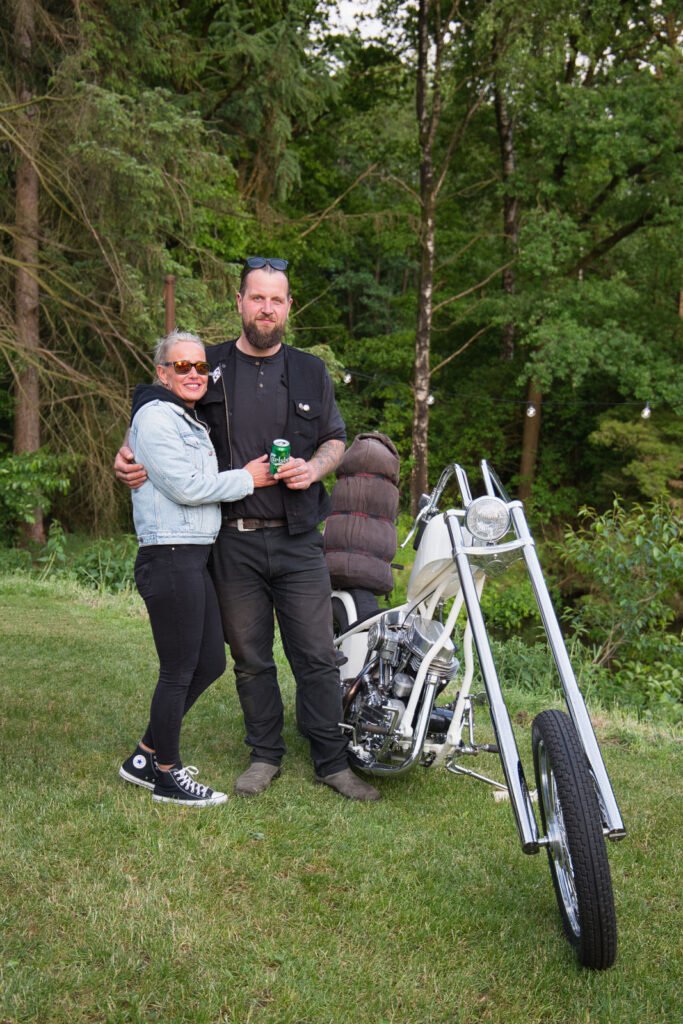 Ein Mann und eine Frau stehen lächelnd neben einem weißen Motorrad im Chopper-Stil auf einer Wiese mit Bäumen im Hintergrund. Der Mann hält ein Getränk in der Hand, und die Frau trägt eine Sonnenbrille und lehnt sich an ihn.