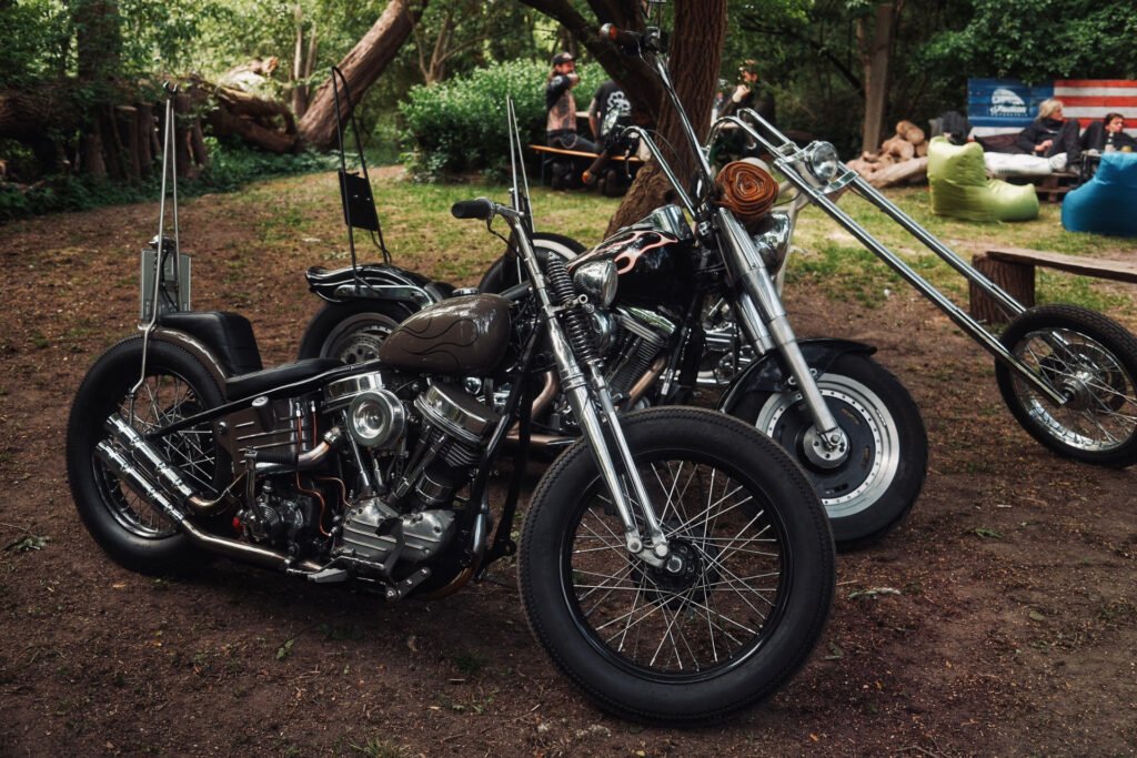 Zwei verchromte Custom-Motorräder mit schwarzen Sitzen sind in einem Waldgebiet unter Bäumen geparkt. Im Hintergrund sitzen Menschen auf Bänken und bunten Sitzsäcken in der Nähe eines Holzzauns.