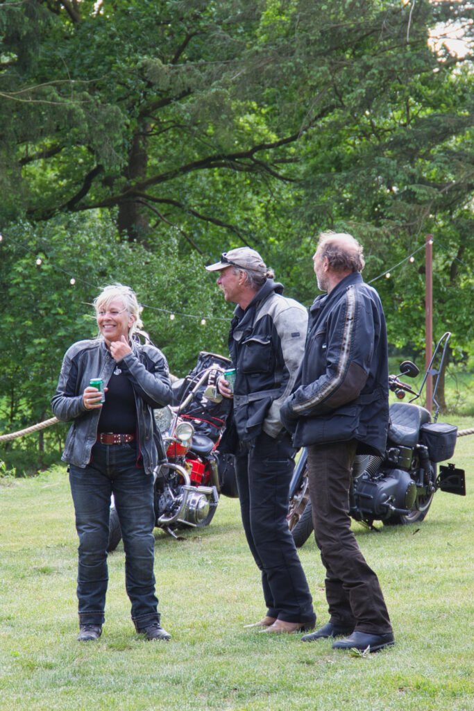 Drei Personen in Motorradkleidung stehen an einem sonnigen Tag im Freien auf einer Wiese, unterhalten sich und lächeln, während hinter ihnen zwei Motorräder geparkt sind und im Hintergrund Bäume zu sehen sind.