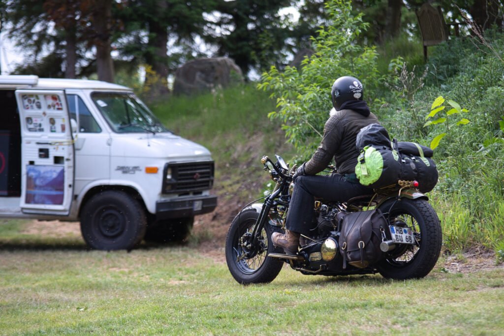Eine Person, die einen Helm und einen Rucksack trägt, fährt mit einem vollgepackten Motorrad auf einer Wiese auf einen weißen Lieferwagen mit Aufklebern zu, der in der Nähe von Bäumen und Grünflächen geparkt ist.