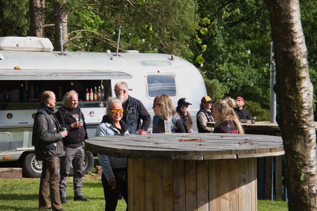 Eine Gruppe von Menschen sitzt im Freien neben einem großen Spulentisch und einem silbernen Anhänger mit Getränken. Einige tragen Jacken und Sonnenbrillen, und Bäume umgeben den Bereich an einem sonnigen Tag.