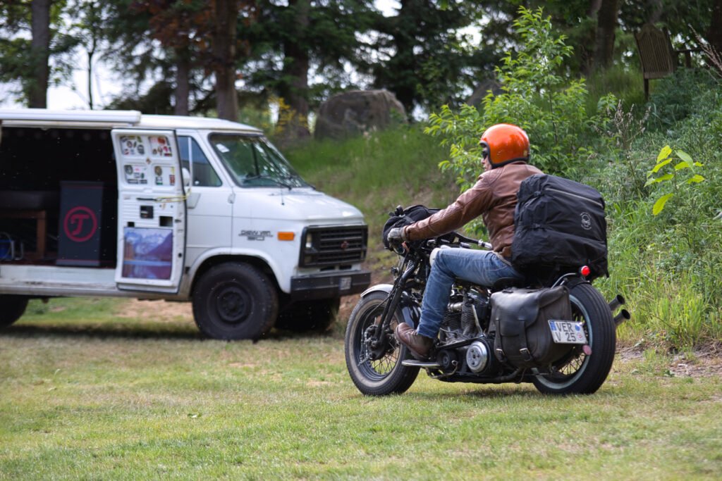 Eine Person, die einen orangefarbenen Helm und eine braune Jacke trägt, fährt auf einem Oldtimer-Motorrad über eine Wiese auf einen weißen Lieferwagen zu, dessen Seitentür geöffnet ist und der in der Nähe von Bäumen und Grünflächen geparkt ist.