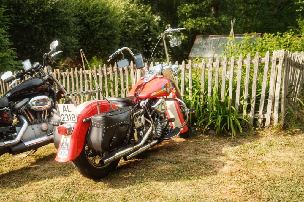 Zwei klassische Motorräder, ein rotes und ein schwarzes, sind an einem sonnigen Tag auf einer Wiese neben einem verwitterten Holzzaun mit Grünpflanzen und einem kleinen Gewächshaus im Hintergrund abgestellt.