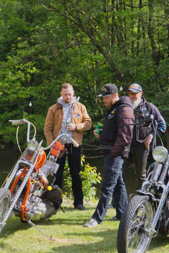 Drei Männer stehen im Freien neben zwei geparkten Motorrädern und unterhalten sich. Sie tragen lässige Jacken und Hüte, im Hintergrund sind Bäume und Grünflächen an einem sonnigen Tag zu sehen.