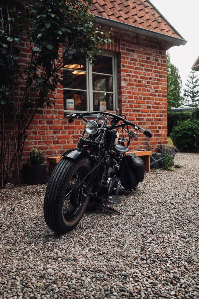 Ein schwarzes Oldtimer-Motorrad mit Satteltaschen steht auf einer Kiesauffahrt vor einem Backsteinhaus mit Ziegeldach und einem Fenster, das warmes Innenlicht reflektiert. In der Nähe befinden sich Sträucher und eine Bank.
