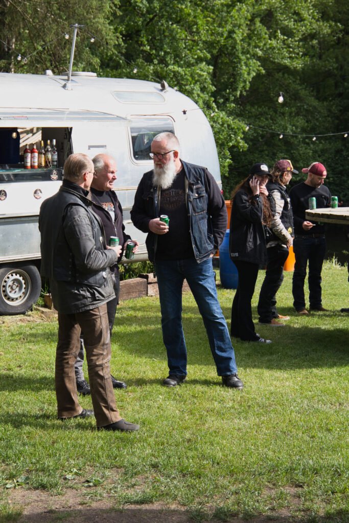 Eine Gruppe von Menschen steht und unterhält sich im Gras neben einem silbernen Imbisswagen mit Getränken, umgeben von grünen Bäumen. Einige halten Dosen, andere unterhalten sich oder telefonieren. Die Szene suggeriert eine zwanglose Zusammenkunft im Freien.