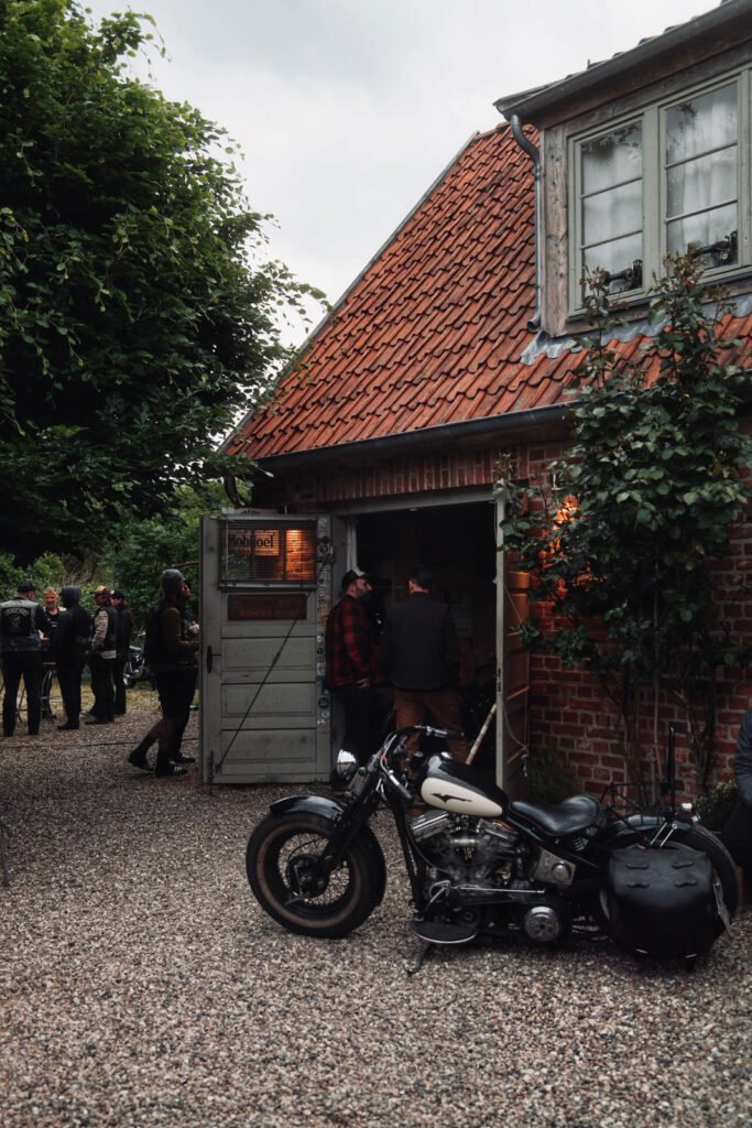 Ein schwarzes Oldtimer-Motorrad ist vor einem Backsteingebäude mit rotem Ziegeldach auf Schotter geparkt. Mehrere Menschen stehen in der Nähe der offenen Tür unter Bäumen, und aus dem Inneren des Gebäudes leuchtet warmes Licht.