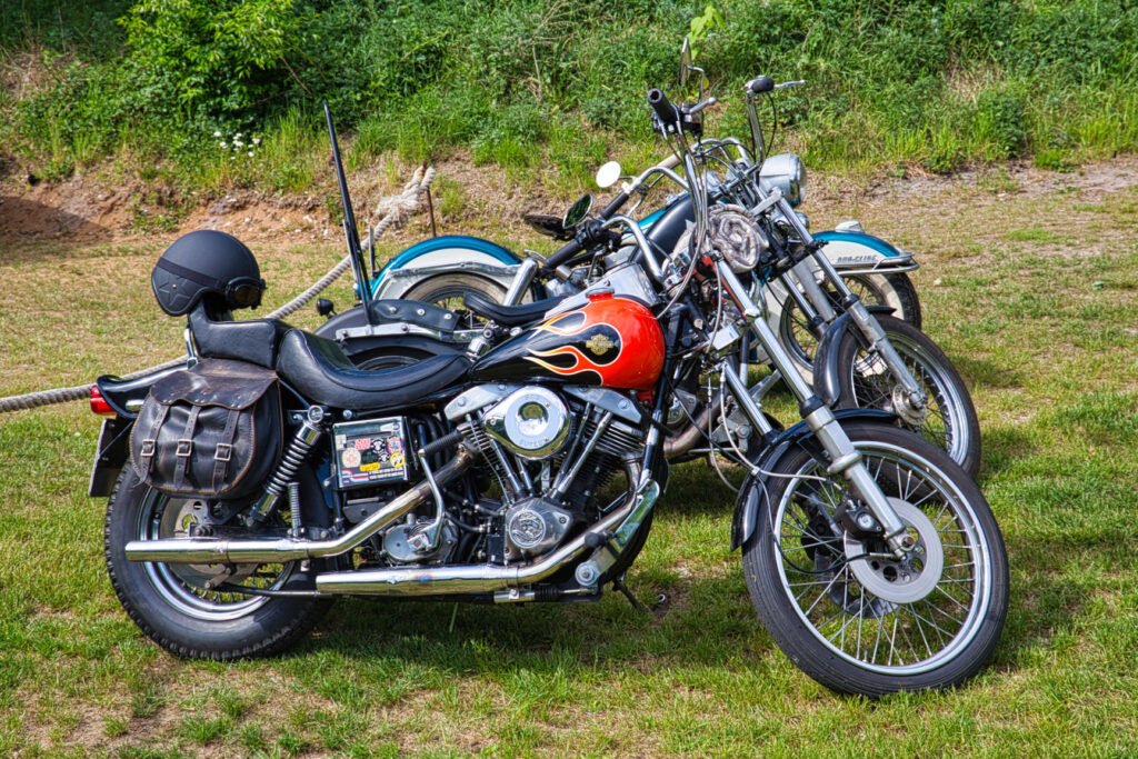 Drei auf einer Wiese geparkte Oldtimer-Motorräder, von denen eines im Vordergrund einen mit Flammen bemalten Benzintank, eine lederne Satteltasche und einen auf dem Sitz ruhenden schwarzen Helm aufweist. Grünes Laub und Schmutz im Hintergrund.