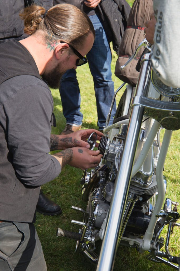 Ein Mann mit Tätowierungen und Sonnenbrille hockt im Freien und arbeitet am Motor eines Motorrads, während mehrere andere Personen in der Nähe im Gras stehen.