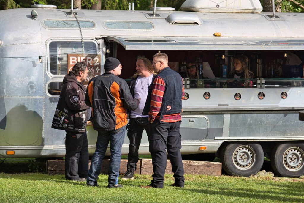 Vier Männer stehen und unterhalten sich vor einem silbernen Imbisswagen auf einer Wiese. Im Inneren des Wagens sind zwei Personen zu sehen, die Getränke servieren. Die Männer tragen legere Outdoor-Kleidung, einige mit Aufnähern auf ihren Jacken.