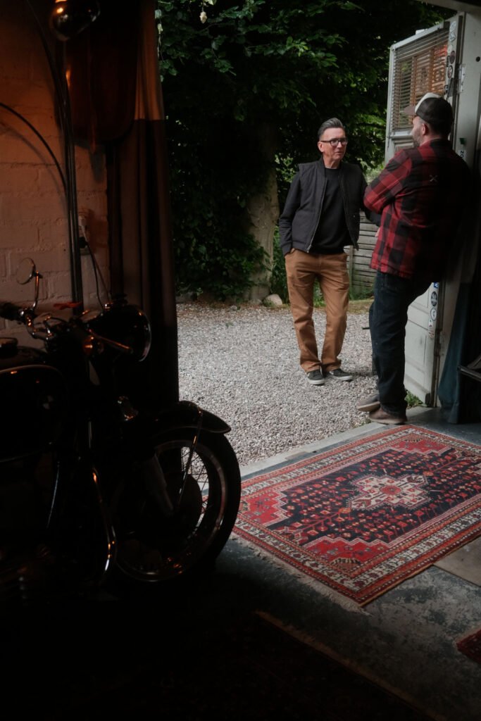 Zwei Männer stehen in einer Tür, die nach draußen führt, und unterhalten sich; der eine trägt eine schwarze Jacke und hellbraune Hosen, der andere ein rotkariertes Hemd und Jeans. Im Inneren sind ein gemusterter Teppich und ein Teil eines Motorrads zu sehen.