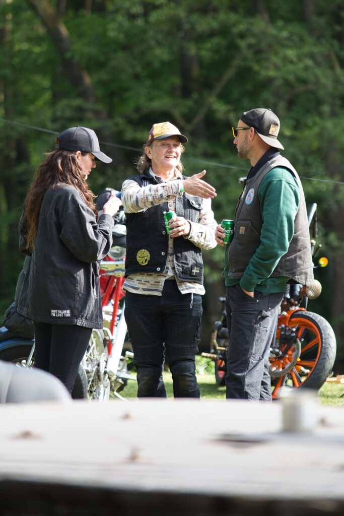 Drei Personen stehen draußen neben Motorrädern, unterhalten sich und halten Getränke in der Hand. Eine Person gestikuliert mit der Hand und lächelt. Die Gruppe ist leger mit Jacken und Hüten gekleidet, mit Bäumen im Hintergrund.