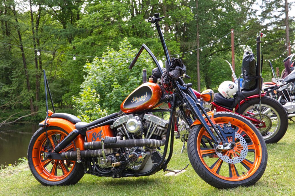 Ein orange-schwarzes Motorrad mit hohem Lenker ist auf einer Wiese in der Nähe eines Teichs geparkt, umgeben von Bäumen und anderen geparkten Motorrädern im Hintergrund.