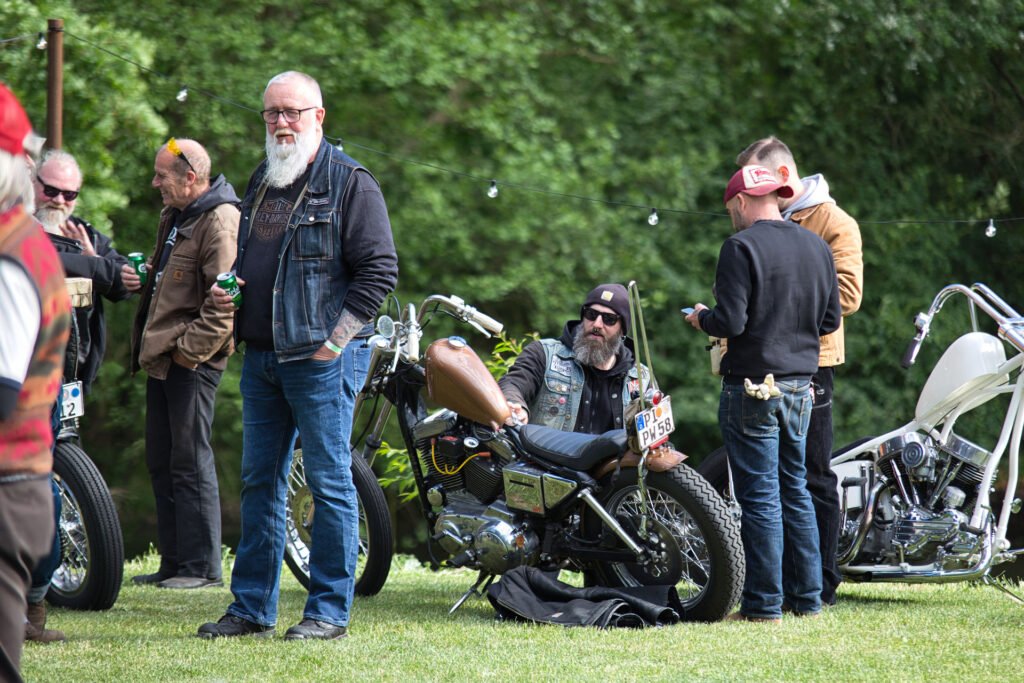 Eine Gruppe von Männern, einige mit Bärten und Tätowierungen, versammelt sich auf einer Wiese neben alten Motorrädern, plaudert und entspannt. Ein Mann sitzt auf einem Motorrad, während andere in der Nähe stehen. Im Hintergrund sind Bäume und Lichterketten zu sehen.